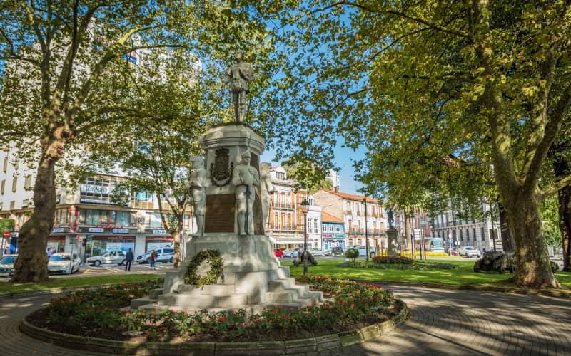 Monumento a Pedro Menéndez de Avilés situado no Parque del Muelle, constituído por uma base escultórica de pedra com figuras e relevos, rodeado de jardins e árvores de grande porte. Ao fundo, edifícios urbanos e uma rua com trânsito num dia de sol e céu limpo.