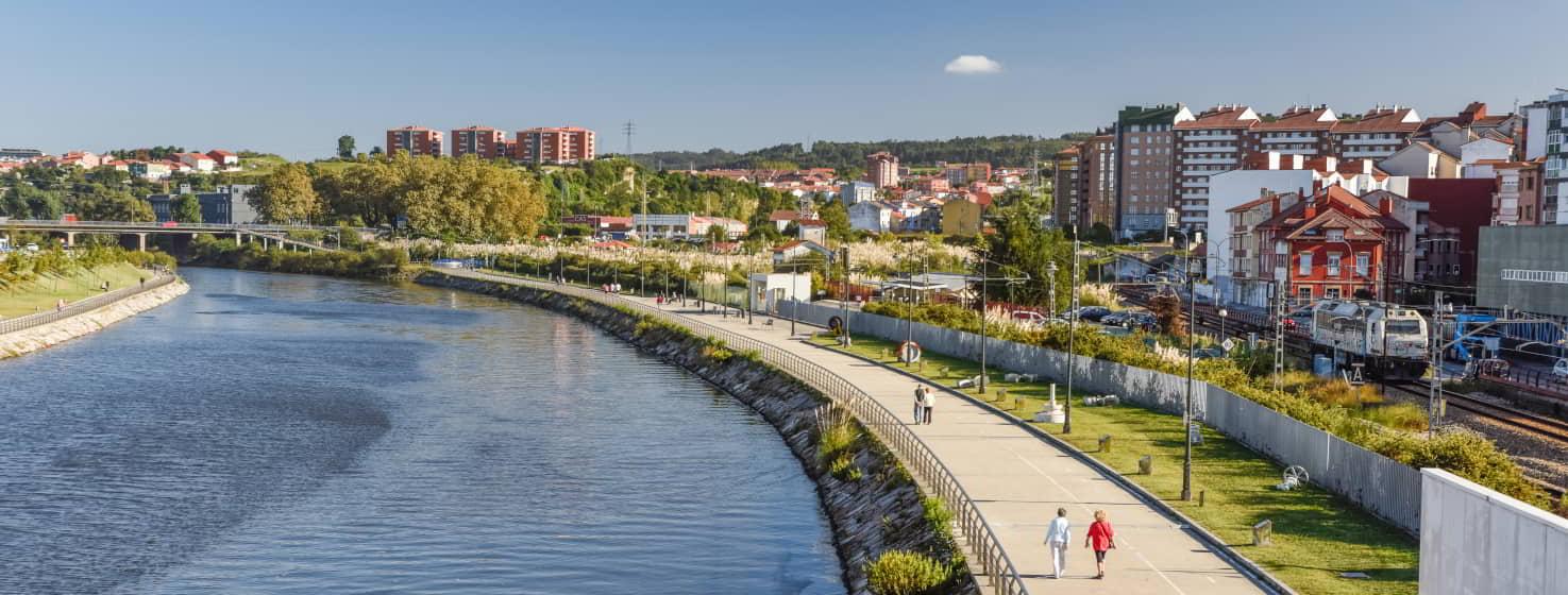 Vista do Paseo de la Ría em Avilés, com um caminho pedonal ao longo da água, ladeado por árvores e jardins. Ao fundo, podem ver-se edifícios residenciais e um comboio a circular junto aos carris, sob um céu limpo.