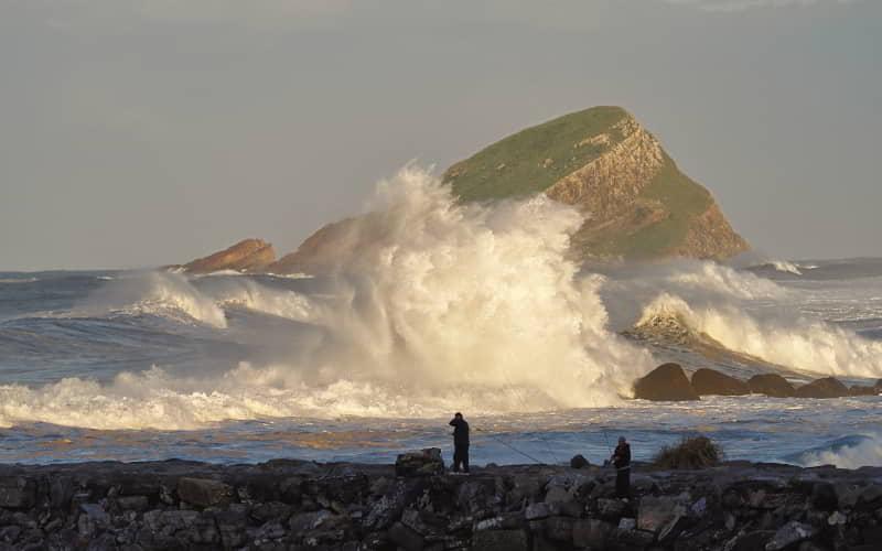 Ola rompiendo con fuerza sobre las rocas en la costa de San Esteban, en Muros de Nalón. En primer plano, dos pescadores lanzan sus cañas desde el espigón, mientras al fondo se alza un islote cubierto de vegetación bajo luz de tarde.