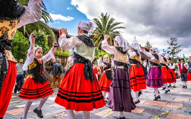 Grupo de pessoas a dançar na rua com trajes tradicionais asturianos durante o Festival de Bollo em Avilés. As dançarinas, vestidas com saias vermelhas e corpetes bordados, levantam os braços ao ritmo da música, rodeadas de palmeiras e do público sob um céu parcialmente nublado.