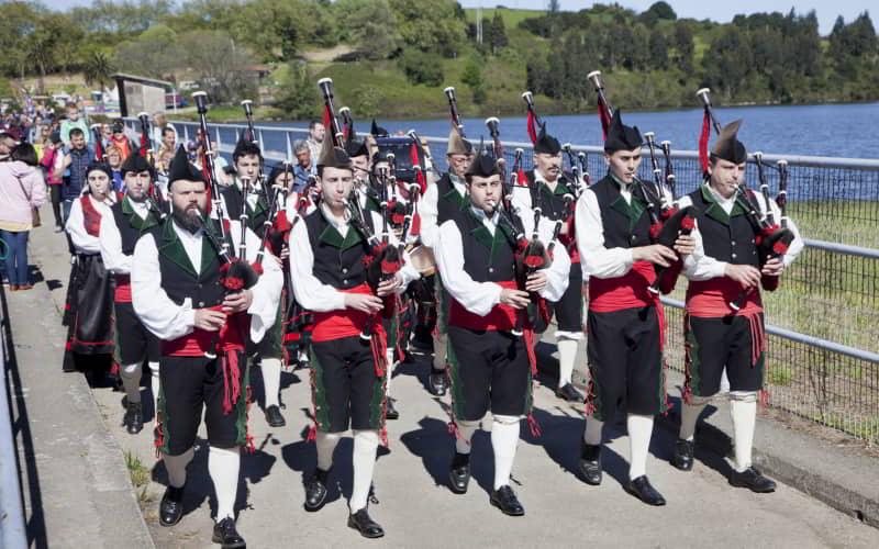 Desfile de gaiteiros vestidos com trajes tradicionais asturianos durante a Jira al Embalse de Trasona, em Corvera de Astúrias. Os músicos avançam ao longo de um passeio junto à água enquanto um grupo de pessoas os segue, num dia de sol.
