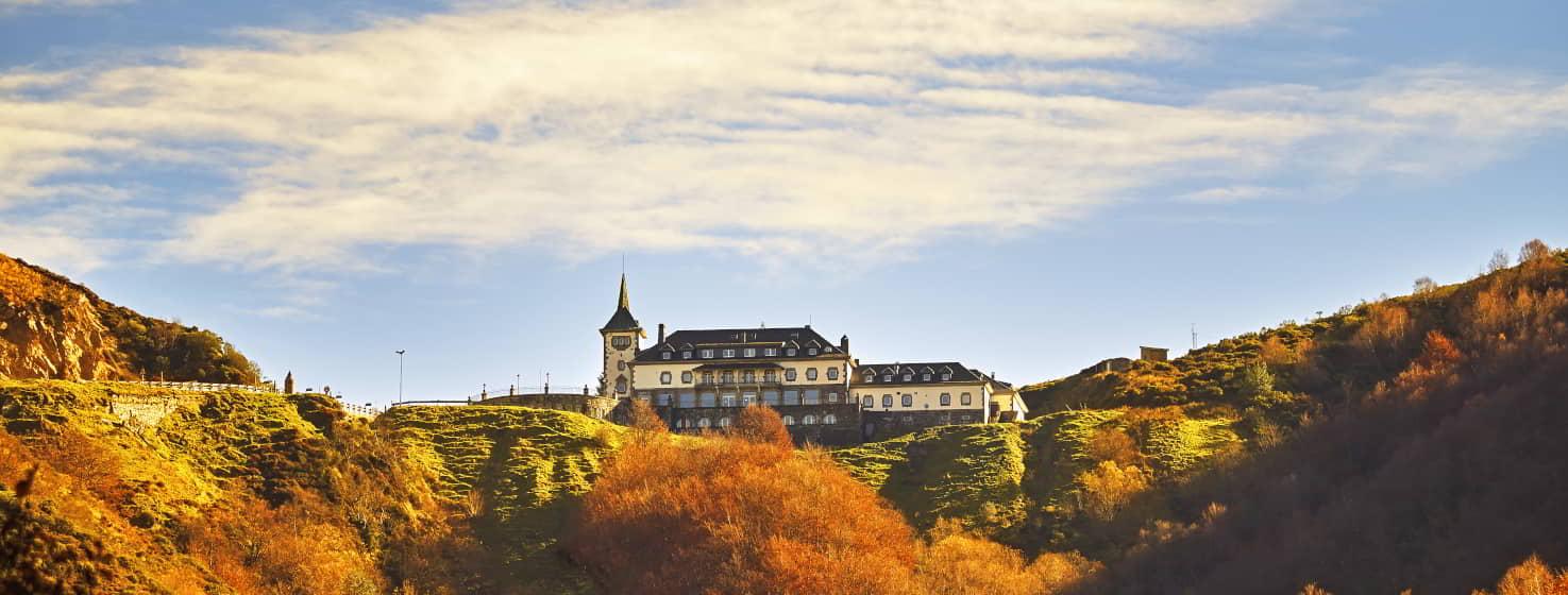 Antiguo Parador de Pajares en Lena, rodeado de un entorno natural en tonos tierra anaranjados, con el edificio al fondo bajo un cielo parcialmente despejado.