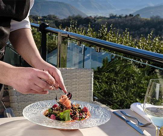 Image of a waiter serving a signature dish on a terrace with the landscape in the background.