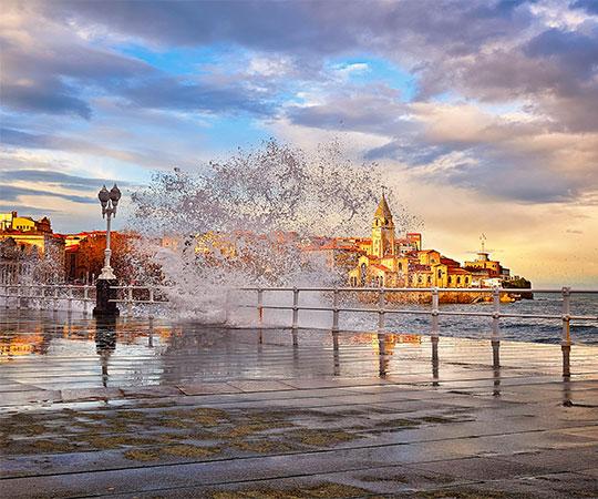 Photo of San Lorenzo Wall Promenade in Gijón/Xixón with waves crashing against the wall and the church of San Pedro in the background