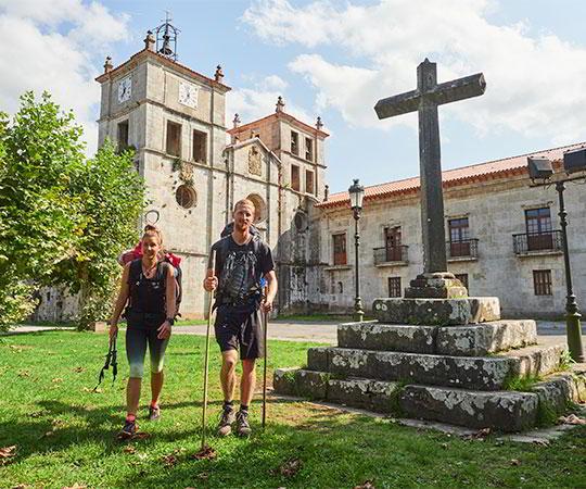 Photo of a couple of pilgrims, on the Primitive Way of St James, in the foreground with the Monastery of San Salvador. Council of Salas.