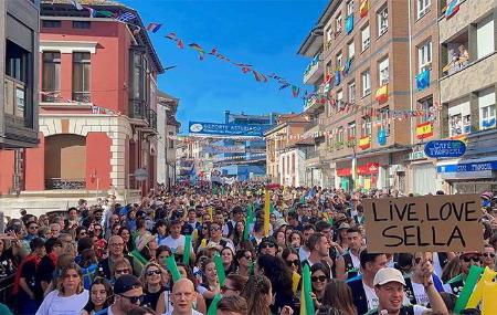 Parade or gathering of people during the Piraguas festival in Arriondas/Les Arriondes, with buildings on either side and garlands of flags.
