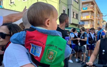 A baby wearing a waistcoat showing the coats of arms of Ribadesella and Cangas de Onís, carried by an adult at the Fiesta del Sella in Asturias.
